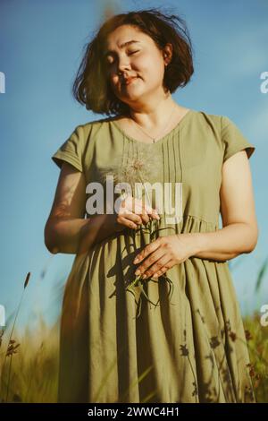 Mature woman holding dandelion and standing in field on sunny day Stock Photo