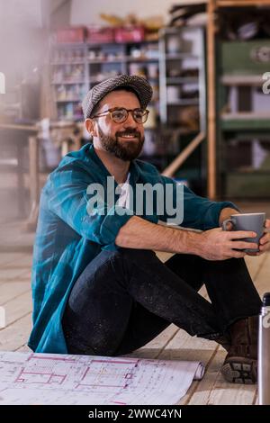 Smiling carpenter sitting by blueprint and holding coffee mug at workshop Stock Photo