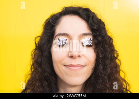 Smiling curly haired woman with butterfly stickers on eyes against yellow background Stock Photo