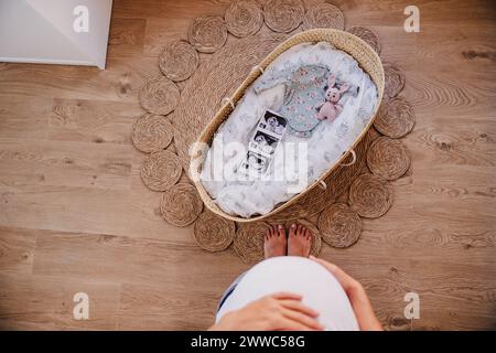 Pregnant woman standing near moses basket on rug at home Stock Photo
