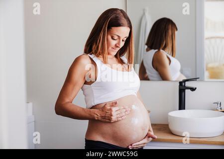 Pregnant woman applying cream on belly near mirror at home Stock Photo