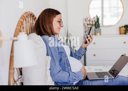 Smiling pregnant freelancer using smart phone near laptop at home office Stock Photo