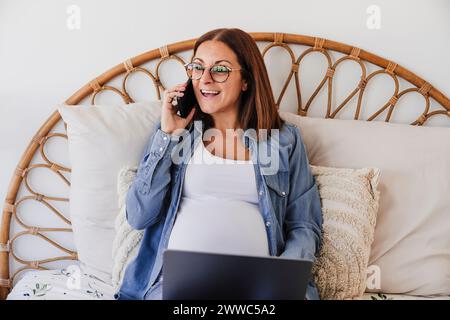 Happy pregnant freelancer talking on smart phone at home office Stock Photo