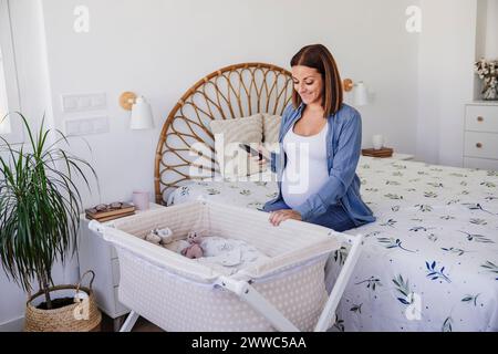 Happy pregnant woman sitting near crib in bedroom at home Stock Photo