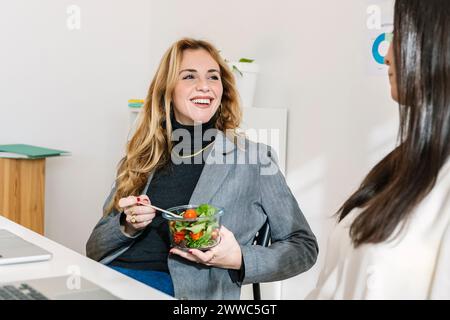 Happy businesswoman sitting with bowl of salad near colleague at desk Stock Photo