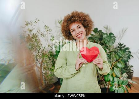 Happy young woman holding red paper heart near plants Stock Photo