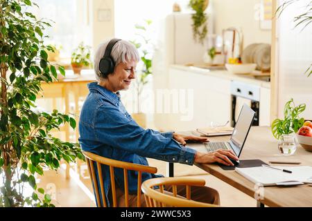 Smiling freelancer wearing wireless headphones using laptop at home office Stock Photo