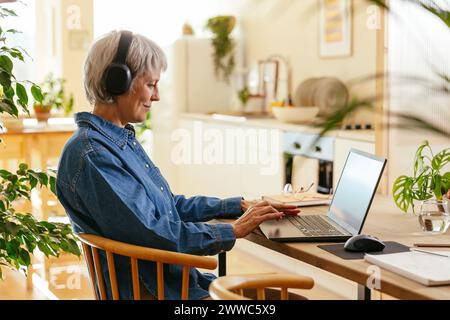 Smiling freelancer wearing wireless headphones and working on laptop at home office Stock Photo