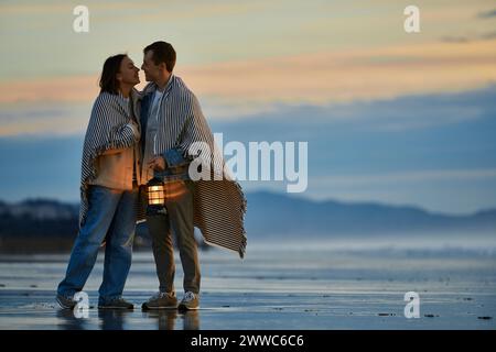 Affectionate couple wrapped in shawl holding lantern at ocean beach Stock Photo