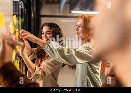 Smiling businesswomen sticking adhesive notes on glass wall in office Stock Photo