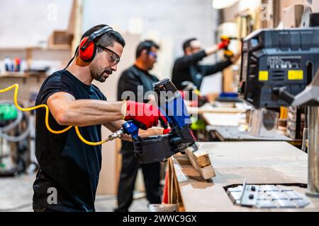 Carpenter using electric hand saw machine on wood with colleagues working in background Stock Photo