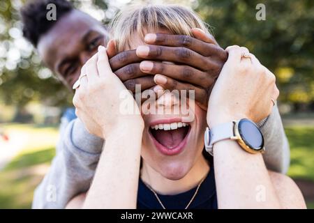 Surprised young woman with eyes covered by friend's hands at park Stock Photo