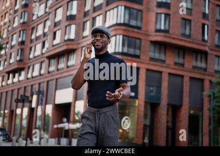 Handsome stylish black man in a cap and black t shirt emotionally engages in a phone conversation and smiles outdoors in the city against a building b Stock Photo