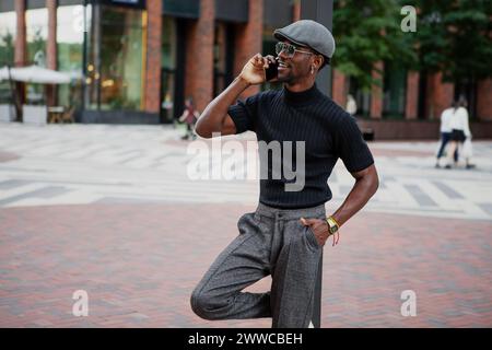 Handsome stylish black man in a cap and black t-shirt emotionally engages in a phone conversation and smiles outdoors in the city against a building b Stock Photo