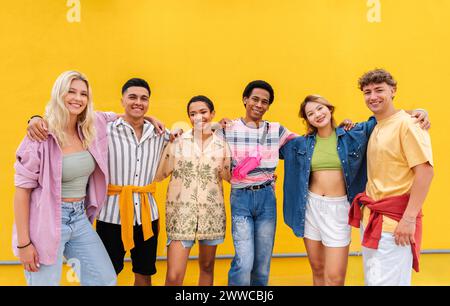 Carefree friends wearing colorful clothing standing in front of yellow wall with arms around Stock Photo