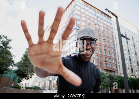stylish, handsome black man outdoors in the city, extends his hand forward, showing gives a high five gesture, wearing a cap and sunglasses, smiling c Stock Photo