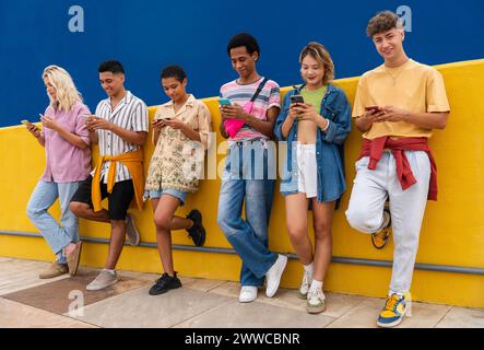 Group of friends leaning against yellow wall using their smartphones Stock Photo