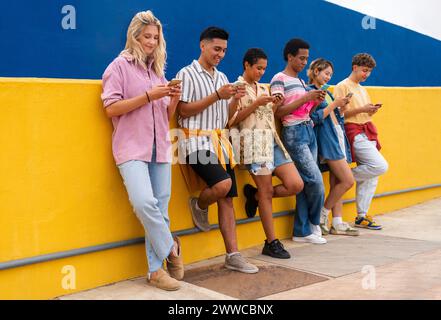 Group of friends leaning against yellow wall using their smartphones Stock Photo
