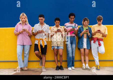 Group of friends leaning against yellow wall using their smartphones Stock Photo