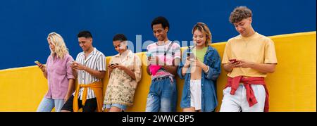Group of friends leaning against yellow wall using their smartphones Stock Photo