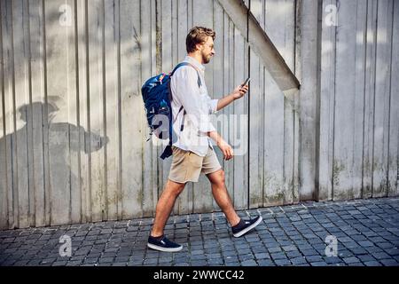 Young man walking with backpack and using smart phone on footpath Stock Photo