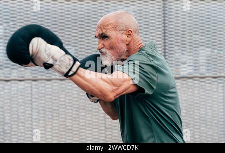 Confident active senior man practicing with boxing gloves in front of fence Stock Photo