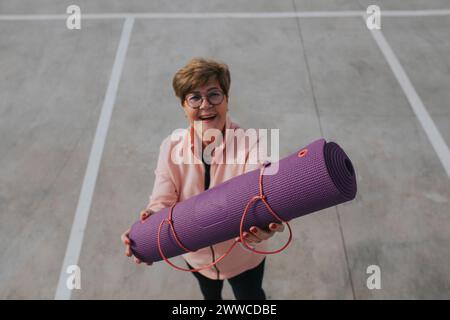 Happy senior woman with exercise mat standing at parking lot Stock Photo