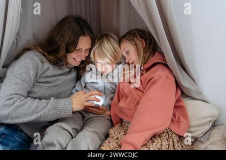 Smiling mother assisting son and daughter using smart phone in canopy at home Stock Photo