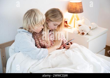 Curious siblings using smart phone sitting on bed at home Stock Photo