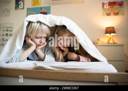Siblings watching smart phone lying down under blanket on bed at home Stock Photo