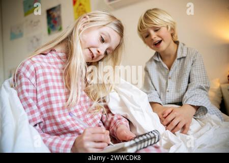 Girl drawing in book sitting with brother on bed at home Stock Photo