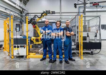 Smiling technician colleagues with arms crossed in front of robotic arm at modern factory Stock Photo