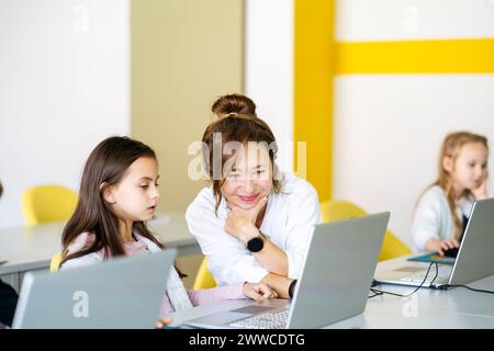 Smiling teacher teaching computer coding on laptop to girl in classroom Stock Photo