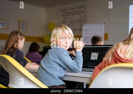 Blond boy looking back over shoulder sitting with laptop at desk in classroom Stock Photo