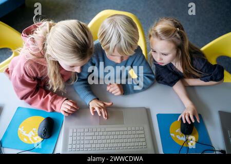 Blond boy sharing laptop with girls learning coding at desk in classroom Stock Photo
