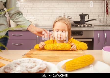 child eats sweet boiled corn in purple kitchen of house. Funny girl at table. Stock Photo