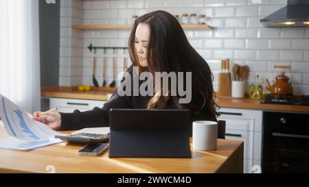Asian woman sit in the kitchen using tablet pay bills, use calculator count expenses, make budget control, manage finances, taking notes. Stock Photo