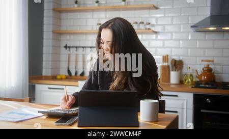 Asian girl sitting in the kitchen using tablet pay bills, use calculator count expenses, make budget control, manage finances, taking notes. Stock Photo