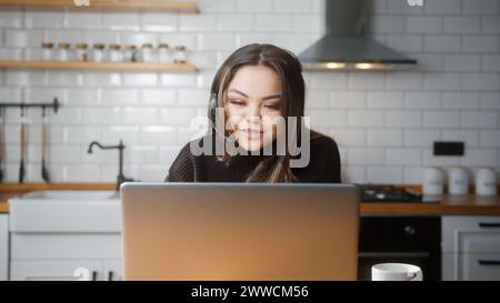 Asian woman wear wireless headset having video conference call on laptop computer talk by webcam in online chat in kitchen at home. Customer support s Stock Photo