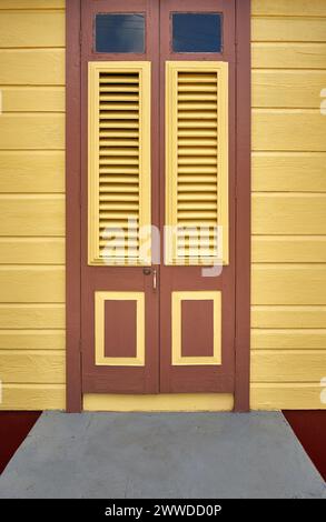 Close-up view of a traditional painted wooden louvered door set against yellow clapboard siding on a Barbadian chattel house. Stock Photo