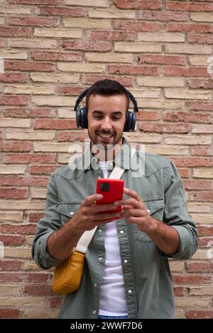 Handsome millennial man using mobile leaning against brick wall. People addicted technology.  Stock Photo
