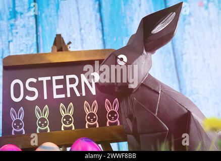 Augsburg, Bavaria, Germany - 23 March 2024: Easter symbol image, board with colorful Easter bunnies and the inscription: Easter, next to an Easter bunny figure. PHOTOMONTAGE *** Ostern Symbolbild, Tafel mit bunten Osterhasen und der Aufschrift: Ostern, neben einer Osterhasenfigur. FOTOMONTAGE Stock Photo