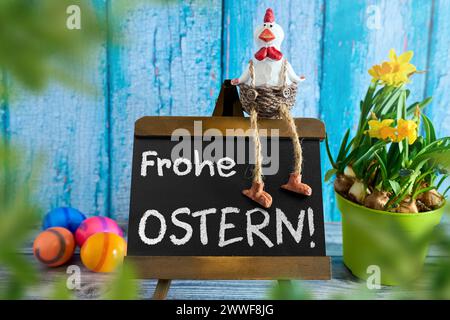 Augsburg, Bavaria, Germany - March 23, 2024: Rooster sits on a plaque with the inscription: Happy Easter next to colorful Easter eggs and daffodils. PHOTOMONTAGE *** Hahn sitzt auf einer Tafel mit Aufschrift: Frohe Ostern neben bunten Ostereiern und Narzissen. FOTOMONTAGE Stock Photo