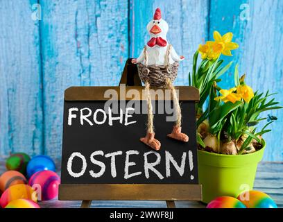 Augsburg, Bavaria, Germany - March 23, 2024: Rooster sits on a plaque with the inscription: Happy Easter next to colorful Easter eggs and daffodils. PHOTOMONTAGE *** Hahn sitzt auf einer Tafel mit Aufschrift: Frohe Ostern neben bunten Ostereiern und Narzissen. FOTOMONTAGE Stock Photo