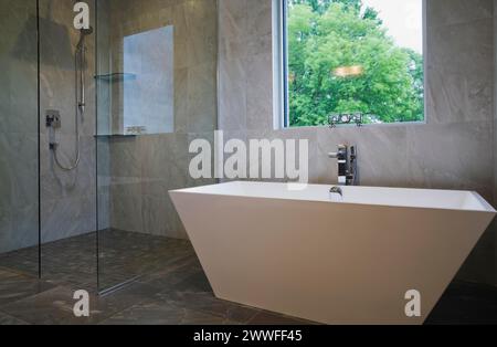 Clear glass shower stall and white freestanding vessel shaped bathtub in bathroom with grey ceramic tile floor and walls on ground floor inside Stock Photo