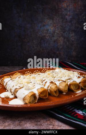 Fried potato tacos with cream and cheese in a Mexican mud dish. Tacos dorados, Mexican food. Stock Photo