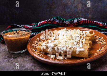 Fried potato tacos with cream and cheese in a Mexican mud dish. Tacos dorados, Mexican food. Stock Photo