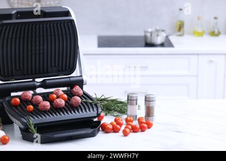 Electric grill with meatballs, tomatoes, spices and vegetables on white marble table in kitchen. Space for text Stock Photo