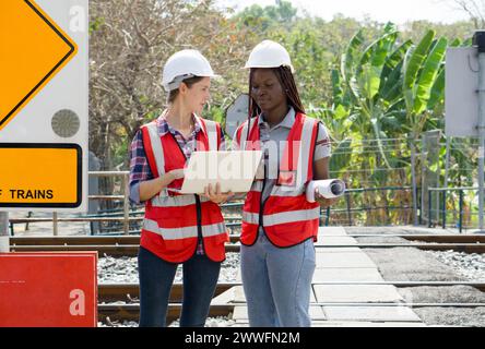 Two individual engaged in a discussion at a railway site, wearing safety equipment, including hard hats and high-visibility vests holding a laptop com Stock Photo