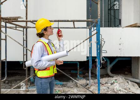 Young female engineer in a construction helmet with a floor plan holding Walkie-Talkie while looking at the progress of real estate projects. Stock Photo